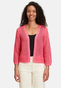 Cardigan en crochet rose avec un tissage ouvert, à manches longues et à coupe décontractée. Porté sur un haut noir, il affiche un col en V et des poignets côtelés.