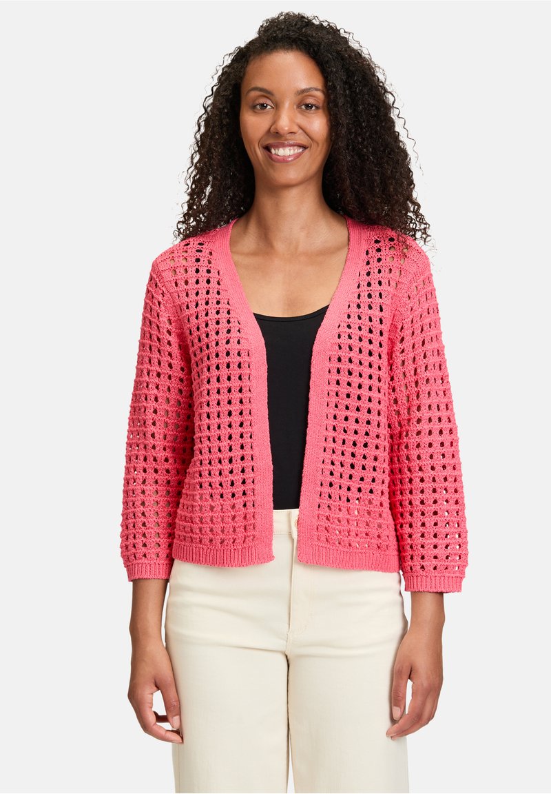 Cardigan en crochet rose avec un tissage ouvert, à manches longues et à coupe décontractée. Porté sur un haut noir, il affiche un col en V et des poignets côtelés.