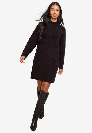 Schwarzes Strickkleid mit gerippter Textur und Zopfmuster, ausgestattet mit hohem Kragen und langen Ärmeln, kombiniert mit kniehohen schwarzen Stiefeln.