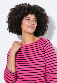 Top a maniche lunghe a righe rosa e nere con scollatura a barca e polsini a coste. Il modello adotta una posa rilassata, mettendo in mostra la texture del tessuto.
