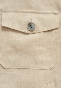 Beige stof med en struktureret vævning har en brystlomme, som er lukket med en sølvfarvet fire-huls knap, syet med lige linjer.