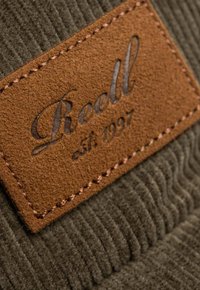 Braunes Lederetikett mit geprägtem "Reell est. 1997" auf grünem Cordstoff-Hintergrund, strukturiert mit vertikalen Rillen und kontrastierenden Nähten.