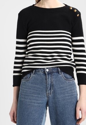 Frau trägt einen schwarz-weiß gestreiften Pullover mit goldenen Knöpfen auf der Schulter, kombiniert mit hoch taillierten blauen Jeans.