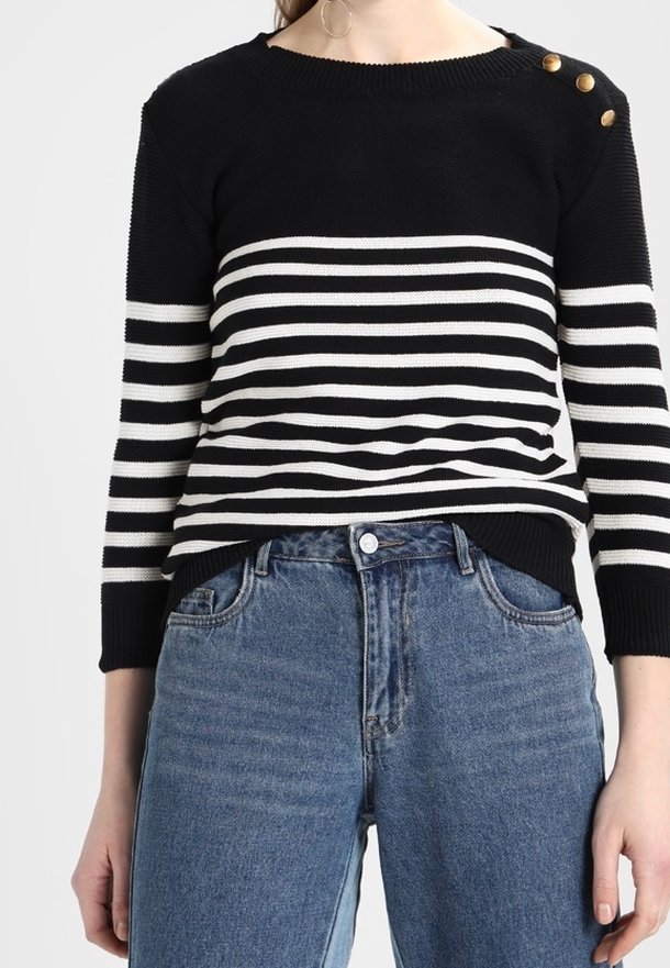 Femme portant un pull à rayures noires et blanches avec des boutons dorés sur l'épaule, associé à un jean bleu taille haute.