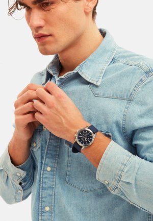 Ung mand i lys denimskjorte, der knapper skjorten, iført armbåndsur med mørkeblå rem og sølvfarvet ramme på venstre håndled.