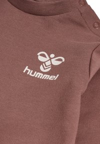 Brauner, texturierter Stoff mit rundem Ausschnitt. Verfügt über Druckknöpfe an der Schulter und ein weißes Logo einer stilisierten Biene mit dem Text "hummel."