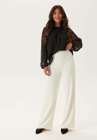 Blusa nera trasparente con maniche lunghe, abbinata a pantaloni bianchi a gamba larga. Il modello indossa scarpe nere con punta a scomparsa. Design semplice ed elegante.