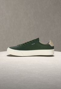 Σκούρο πράσινο χαμηλό αθλητικό παπούτσι με λευκή σόλα και μπεζ ταμπελάκι στη φτέρνα, τοποθετημένο σε ουδέτερη επιφάνεια μπροστά σε γκρίζο φόντο.