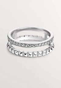 Dos anillos de plata apilados uno sobre el otro. El anillo de arriba presenta pequeños cristales claros, mientras que el anillo de abajo tiene un diseño tachonado con formas de pirámide.