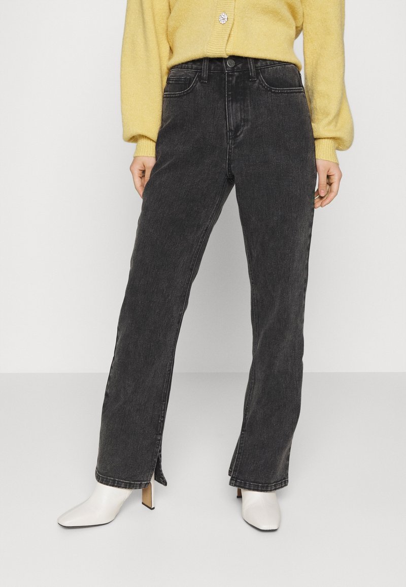 Femme portant un jean noir taille haute à jambes droites avec fentes latérales, un cardigan jaune et des bottines blanches à talons, debout sur un sol gris.