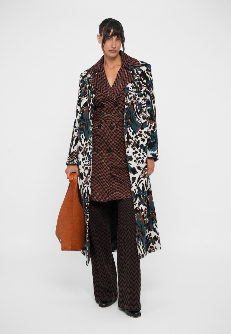 Lange, patroon trenchcoat met dierenprint en levendige kleuren, gedragen over een gestructureerde bruine jurk en wijde broek, met een oranje tas.