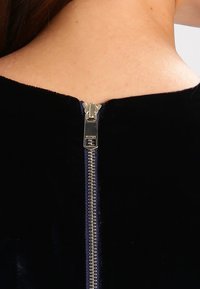 Prenda de terciopelo negro con un cuello alto y un detalle de cremallera plateada en la parte posterior, que presenta un acabado texturizado, suave y un diseño elegante.