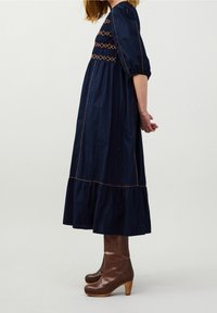 Marineblaues Kleid mit gestuftem Saum, versehen mit orangefarbenen Nähten und einem gerafften Oberteil. Kombiniert mit braunen Plateaustiefeletten.