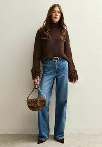 Pull en tricot marron avec des manches à franges, un jean en denim bleu taille haute et un sac à main en cuir texturé imprimé serpent marron. Talons aiguilles marron.