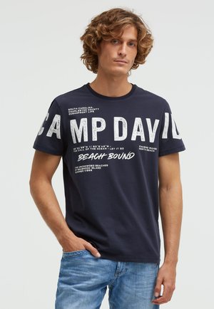 Young man with curly hair wearing a navy blue "Camp David Beach Bound" t-shirt and light blue jeans, standing against a plain background.