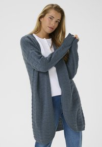 Femme portant un long cardigan en tricot gris sur une chemise blanche et un jean bleu, ajustant sa manche sur un fond neutre.