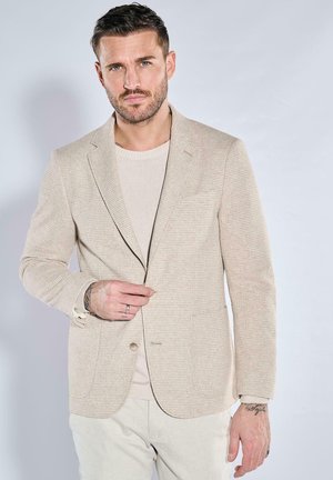 Beige strukturierter Blazer mit einem einzelnen Knopf, Reverskragen und zwei Fronttaschen, über einem hellen Pullover getragen, kombiniert mit hellen Hosen.