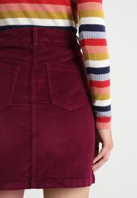 Burgundyröd kordfløjelskjol med bakfickor, hög midja och en texturerad yta. Randig långärmad tröja delvis synlig.