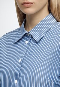 Femme portant une chemise bleue avec des rayures verticales blanches et des boutons blancs, montrant le col et le haut de la poitrine.