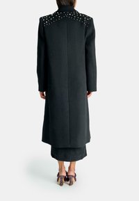Cappotto nero lungo con spalle borchiate, taglio dritto, realizzato in tessuto testurizzato. Presenta una schiena solida ed è indossato sopra una gonna nera e tacchi.