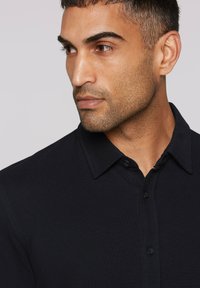Dark navy polo shirt with a textured fabric, button placket, and a classic collar. Close-up of face, with short hair and stubble.