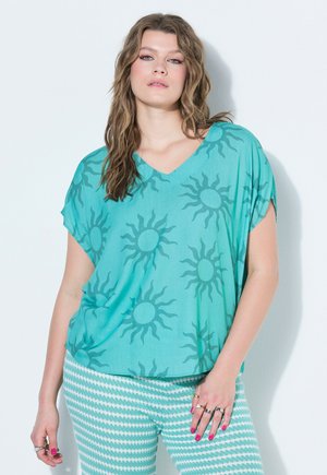 Kvinde med langt bølget hår iført løs t-shirt med teal solmønster og teal-hvide mønstrede bukser, poserer mod en hvid baggrund.