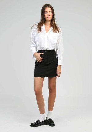 Femme debout portant une chemise blanche à boutons, une jupe noire ornée, des chaussettes blanches et des mocassins noirs sur un fond gris uni.