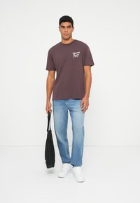 Brun bomull t-skjorte med hvit logo, kombinert med lyseblå denimjeans og hvite sneakers. Holder en svart totebag. Enkel, avslappet passform.
