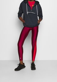 Schwarzer leichter Hoodie mit Fronttasche, kombiniert mit glänzenden roten Leggings mit schwarzen Seitenstreifen und schwarzen Sportschuhen.