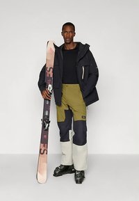 Mann iført svart jakke, svart skjorte, fargeblokkede skibukser og skistøvler som holder et enkelt rosa og svart ski oppreist.