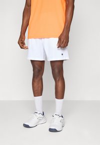 Shorts de sport blancs avec un logo noir, associés à des chaussettes montantes blanches et des chaussures blanches avec des accents bleu marine. Un t-shirt orange est visible.