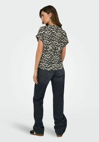 Kortärmad topp med ett svart och crèmefärgat abstrakt mönster, som har en rundad fåll och en lös passform, kombinerad med mörkblå vida jeans.