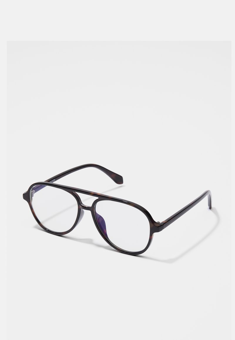 Lunettes aviator en écaille noire avec des verres transparents reposant sur une surface blanche.