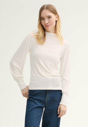Top blanco de manga larga con un cuello alto y cintura ajustada, con mangas ligeramente abullonadas y una textura suave y lisa. Combinado con jeans azules.
