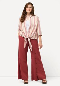 Chemise à boutons rayée rose et blanche nouée à la taille, associée à un pantalon rouge à jambes larges avec des boutons sur le côté. Sandales à plateforme brunes.
