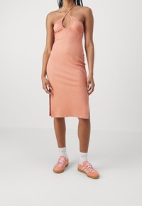 Femme portant une robe halter sans manches, longueur genou, de couleur pêche, accompagnée de chaussettes blanches et de baskets assorties en pêche, sur fond blanc.