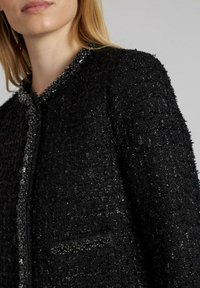 Schwarze texturierte Jacke mit einem mit Perlen verzierten Kragen und Details an der Vorderseite der Taschen, die eine strukturierte Silhouette und Paillettenakzente aufweist.