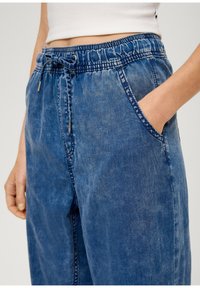 Světle modré džíny s elastickým pasem, šňůrkovým detailem a bočními kapsami. Materiál má měkký, strukturovaný vzhled.