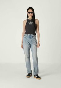 Žena stojí v čiernom bezrukávovom topu s bielym kvetinovým vzorom, svetlomodrých džínsach, čiernych teniskách, so slnečnými okuliarmi a farebným korálkovým náhrdelníkom.