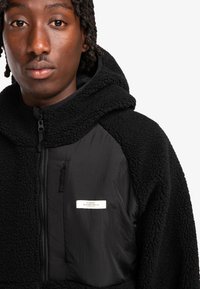 Sudadera con capucha negra de forro polar, con un exterior texturizado, detalles de nailon lisos, bolsillos con cremallera y una etiqueta que presenta "ELEMENT SKATEBOARD CO."