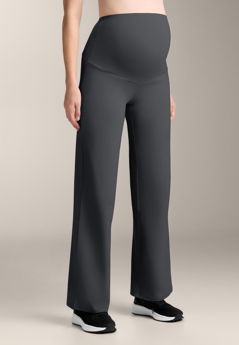 Schwangere Person, die schwarze Umstandshosen und schwarze Sneakers trägt, vor einem neutralen Hintergrund stehend.