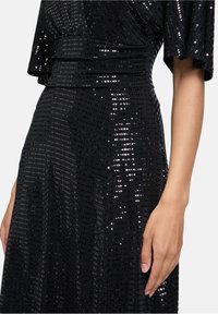 Robe noire à sequins avec une silhouette fluide, dotée d'une surface texturée et de larges manches, mettant en valeur un motif de sequins circulaires.