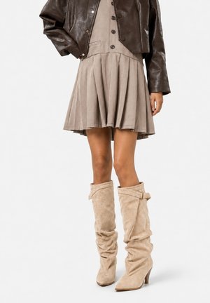 Vrouw draagt een beige geplooide rok, bruine slappe kniehoge laarzen met gespen en een kort donkebruin leren jack.
