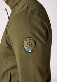 Veste zippée vert olive avec un col haut, ornée d'un patch circulaire brodé avec des avions et des accents bleus, blancs et rouges sur la manche.