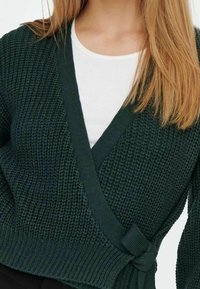 Dunkelgrüner, gestrickter Cardigan mit Wickeldesign und Gürteldetail. Strukturierter Stoff, V-Ausschnitt, über einem weißen Hemd getragen.