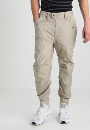 Mann trägt beige wandelbare Cargohose und weiße Sneakers, Hände in den Taschen, steht vor einem schlichten hellen Hintergrund.