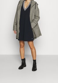 Zelená parka s kapucí, přes námořnické šaty s výstřihem do V a bílými pruhy. Černé kotníkové boty doplňují outfit. Pozorovány jemné textury.