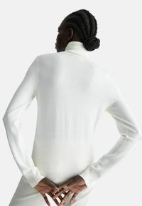 Maglione bianco a collo alto con maniche lunghe, polsini a coste e trama a maglia liscia, che mostra un design semplice e minimalista dal retro.