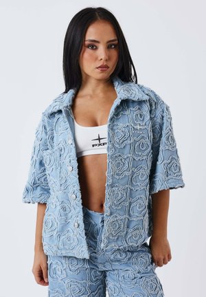 Vrouw met stijl zwart haar draagt een lichtblauwe gestructureerde jas en broek met bloemenpatronen en een witte crop top met zwart logo.