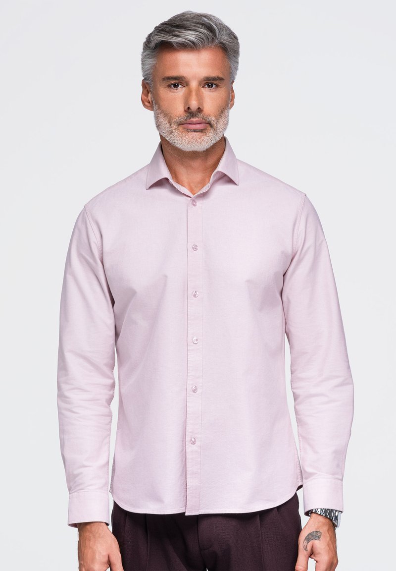 Homme aux cheveux gris et à la barbe, portant une chemise rose clair à boutons et un pantalon foncé, debout devant un fond blanc uni.